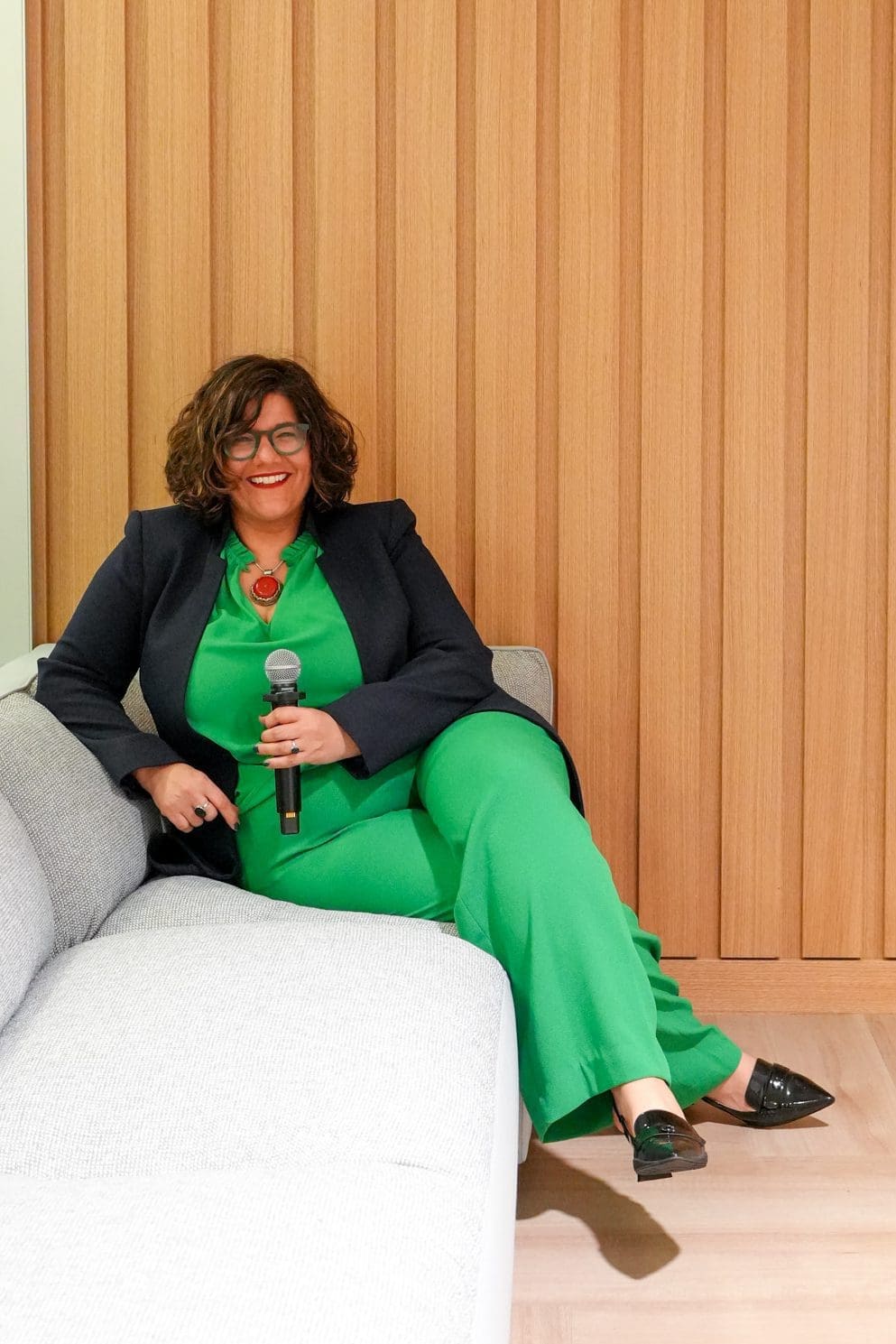 A woman in a green outfit sitting on a gray couch against a wooden wall.