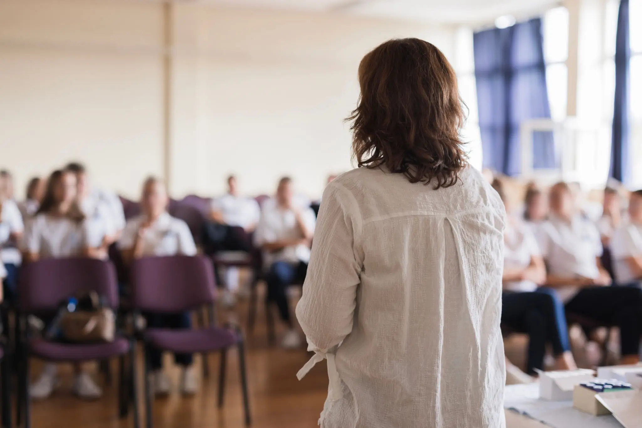 A woman presenting to a seated audience in a bright room.